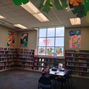 Photograph of the final panel art pieces hung on the wall of the Teen Room. There are three brightly colored paintings hanging above bookshelves and next to large windows.