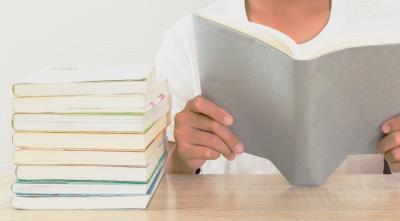 Photograph of a stack of books set on a table. Next to the stack, a person's hands are holding a gray book open.