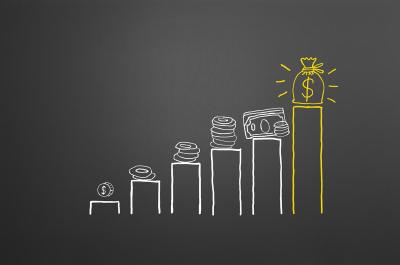 A black chalkboard with a bar graph in white chalk showing increasing amounts of coins, the last bar is the highest and it's yellow with a money bag on top.