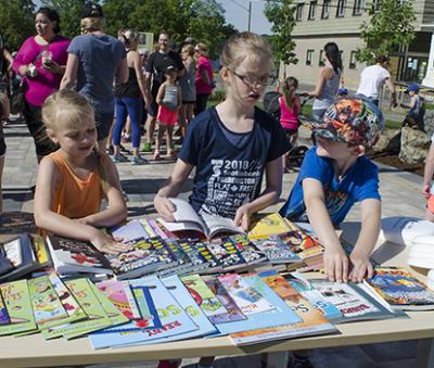 Three young children browsing books on a table at an outdoor event.