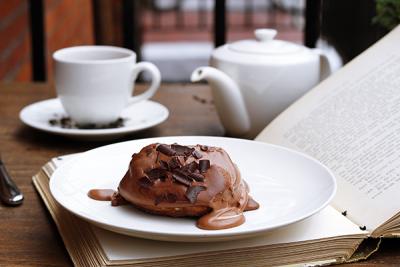 A small chocolate cake sits on a plate on top of an open book. A tea cup and tea pot are in the background.