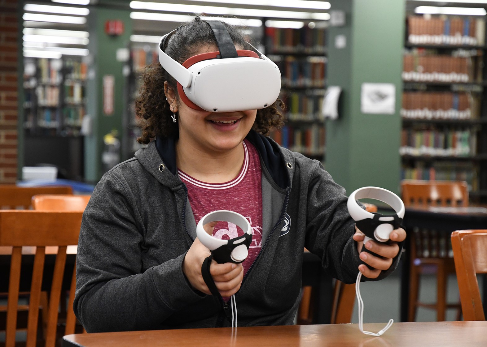 Photograph shows a student with a VR headset and hand controllers. They are sitting at a desk inside of the library.
